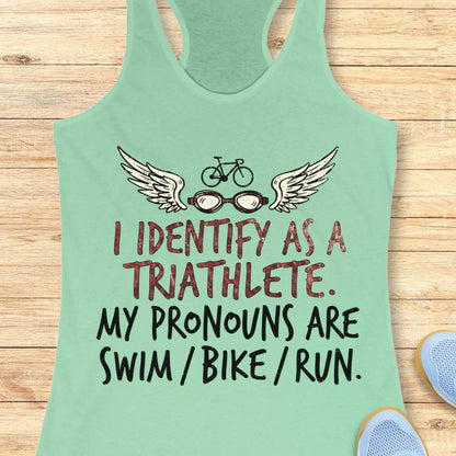 mint racerback tank top displayed on a light wood background, featuring a humorous triathlon-themed design with a bicycle above a pair of winged swim goggles. The graphic includes distressed red and black lettering that reads: “I IDENTIFY AS A TRIATHLETE. MY PRONOUNS ARE SWIM / BIKE / RUN.” Partial blue running shoes are visible in the bottom corners of the image.