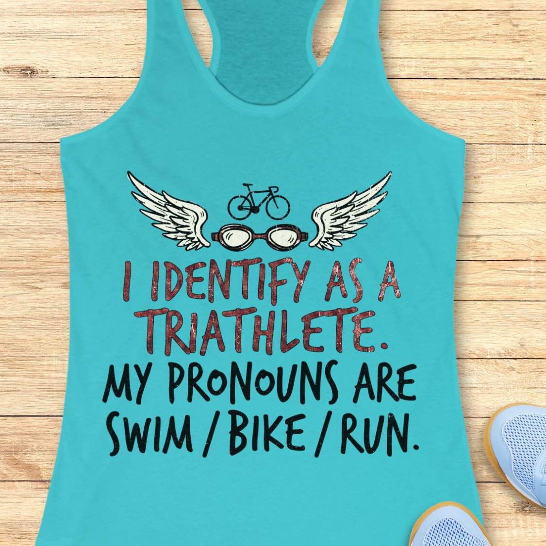 blue racerback tank top displayed on a light wood background, featuring a humorous triathlon-themed design with a bicycle above a pair of winged swim goggles. The graphic includes distressed red and black lettering that reads: “I IDENTIFY AS A TRIATHLETE. MY PRONOUNS ARE SWIM / BIKE / RUN.” Partial blue running shoes are visible in the bottom corners of the image.