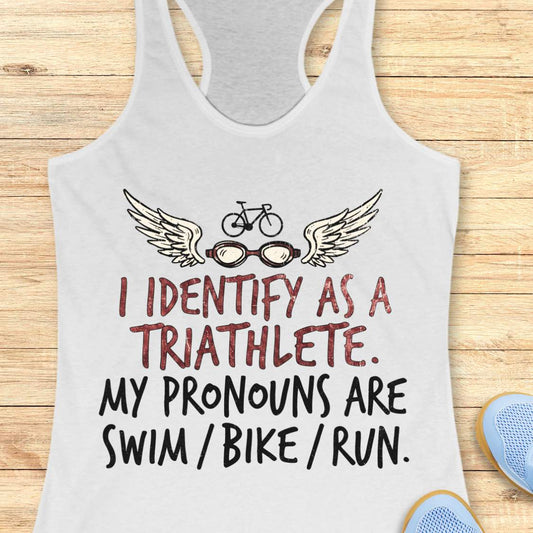 White racerback tank top displayed on a light wood background, featuring a humorous triathlon-themed design with a bicycle above a pair of winged swim goggles. The graphic includes distressed red and black lettering that reads: “I IDENTIFY AS A TRIATHLETE. MY PRONOUNS ARE SWIM / BIKE / RUN.” Partial blue running shoes are visible in the bottom corners of the image.