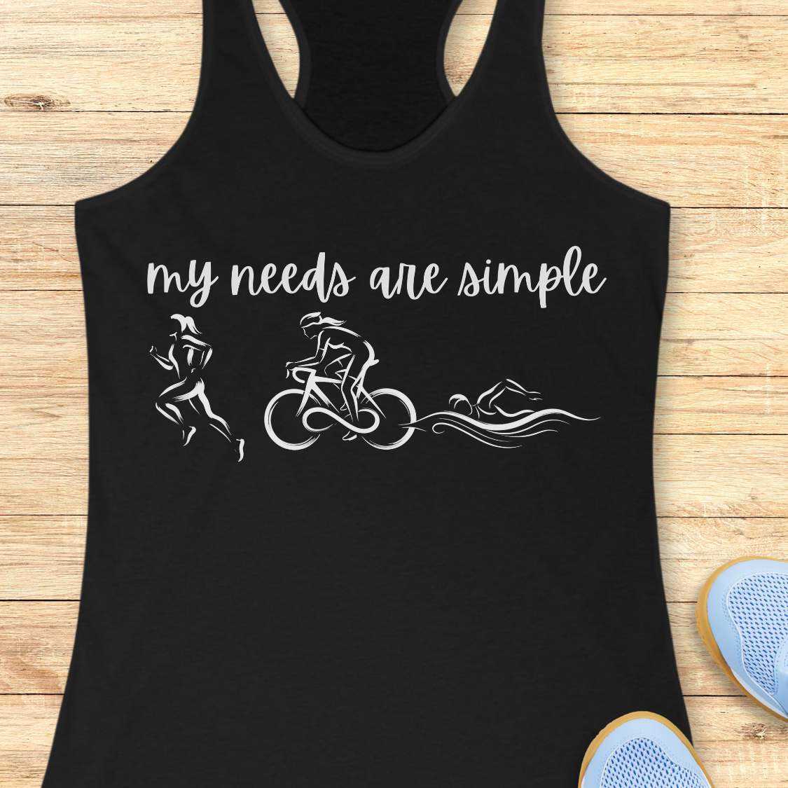 black racerback tank top displayed on a light wood background, featuring a minimalist triathlon-themed design. The graphic includes the phrase “my needs are simple” in black script lettering above line-art illustrations of a runner, cyclist, and swimmer, representing the three disciplines of triathlon. Partial blue running shoes are visible in the bottom corners of the image.