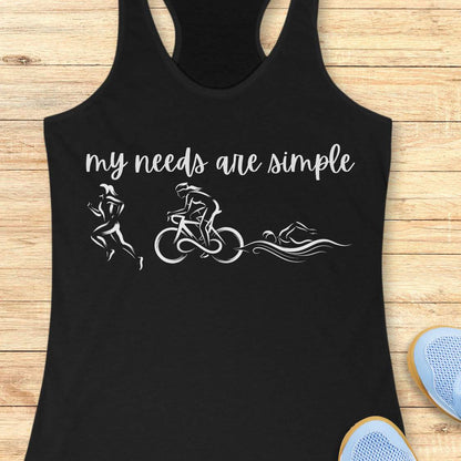 black racerback tank top displayed on a light wood background, featuring a minimalist triathlon-themed design. The graphic includes the phrase “my needs are simple” in black script lettering above line-art illustrations of a runner, cyclist, and swimmer, representing the three disciplines of triathlon. Partial blue running shoes are visible in the bottom corners of the image.