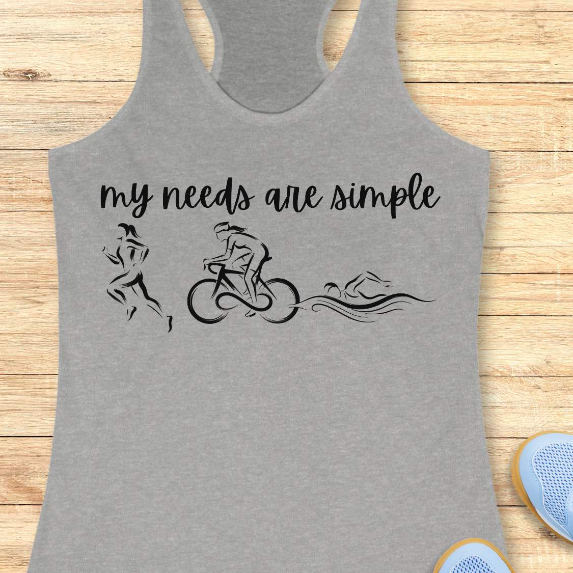 grey racerback tank top displayed on a light wood background, featuring a minimalist triathlon-themed design. The graphic includes the phrase “my needs are simple” in black script lettering above line-art illustrations of a runner, cyclist, and swimmer, representing the three disciplines of triathlon. Partial blue running shoes are visible in the bottom corners of the image.