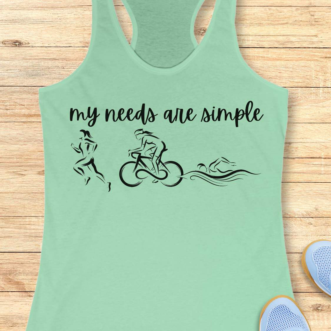 mint racerback tank top displayed on a light wood background, featuring a minimalist triathlon-themed design. The graphic includes the phrase “my needs are simple” in black script lettering above line-art illustrations of a runner, cyclist, and swimmer, representing the three disciplines of triathlon. Partial blue running shoes are visible in the bottom corners of the image.