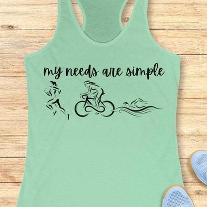 mint racerback tank top displayed on a light wood background, featuring a minimalist triathlon-themed design. The graphic includes the phrase “my needs are simple” in black script lettering above line-art illustrations of a runner, cyclist, and swimmer, representing the three disciplines of triathlon. Partial blue running shoes are visible in the bottom corners of the image.