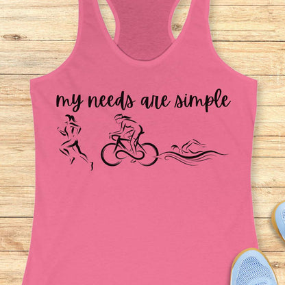 A pink racerback tank top laid flat on a light wood background, featuring the black printed text 'my needs are simple' above minimalist silhouettes of a runner, cyclist, and swimmer.