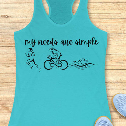 Tahiti blue racerback tank top displayed on a light wood background, featuring a minimalist triathlon-themed design. The graphic includes the phrase “my needs are simple” in black script lettering above line-art illustrations of a runner, cyclist, and swimmer, representing the three disciplines of triathlon. Partial blue running shoes are visible in the bottom corners of the image.