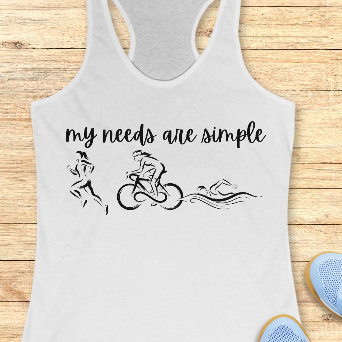 white racerback tank top displayed on a light wood background, featuring a minimalist triathlon-themed design. The graphic includes the phrase “my needs are simple” in black script lettering above line-art illustrations of a runner, cyclist, and swimmer, representing the three disciplines of triathlon. Partial blue running shoes are visible in the bottom corners of the image.
