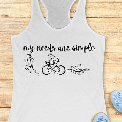 white racerback tank top displayed on a light wood background, featuring a minimalist triathlon-themed design. The graphic includes the phrase “my needs are simple” in black script lettering above line-art illustrations of a runner, cyclist, and swimmer, representing the three disciplines of triathlon. Partial blue running shoes are visible in the bottom corners of the image.