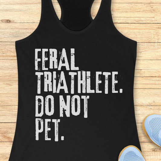 Black racerback tank top displayed on a light wood background, featuring bold distressed white lettering that reads: “FERAL TRIATHLETE. DO NOT PET.” The minimalist design uses large vintage-style typography for a humorous triathlon-themed statement. Partial blue running shoes are visible in the bottom corners of the image.