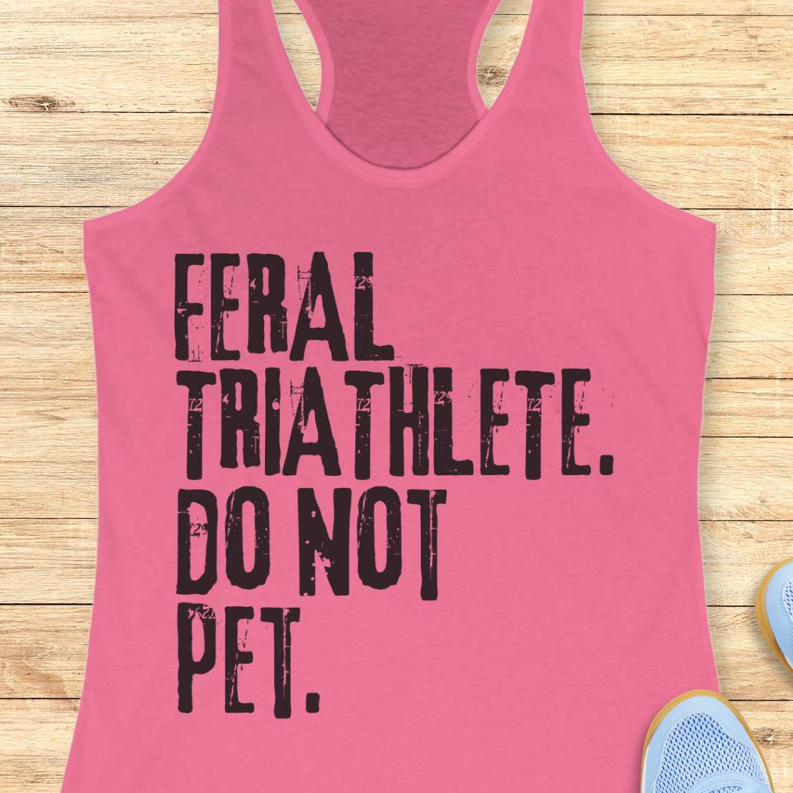 pink racerback tank top displayed on a light wood background, featuring bold distressed white lettering that reads: “FERAL TRIATHLETE. DO NOT PET.” The minimalist design uses large vintage-style typography for a humorous triathlon-themed statement. Partial blue running shoes are visible in the bottom corners of the image.