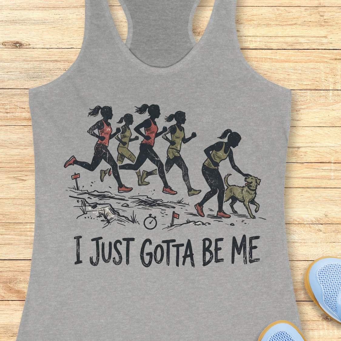 Heather gray racerback tank top featuring a distressed vintage-style illustration of five runners on a trail. Four runners continue the race while one runner stops to pet a friendly dog beside the path. Small trail markers and a stopwatch appear in the scene. The design includes the phrase: “I JUST GOTTA BE ME.” The artwork uses muted coral, olive green, and black tones, celebrating runners who prioritize joy and dogs over competition.