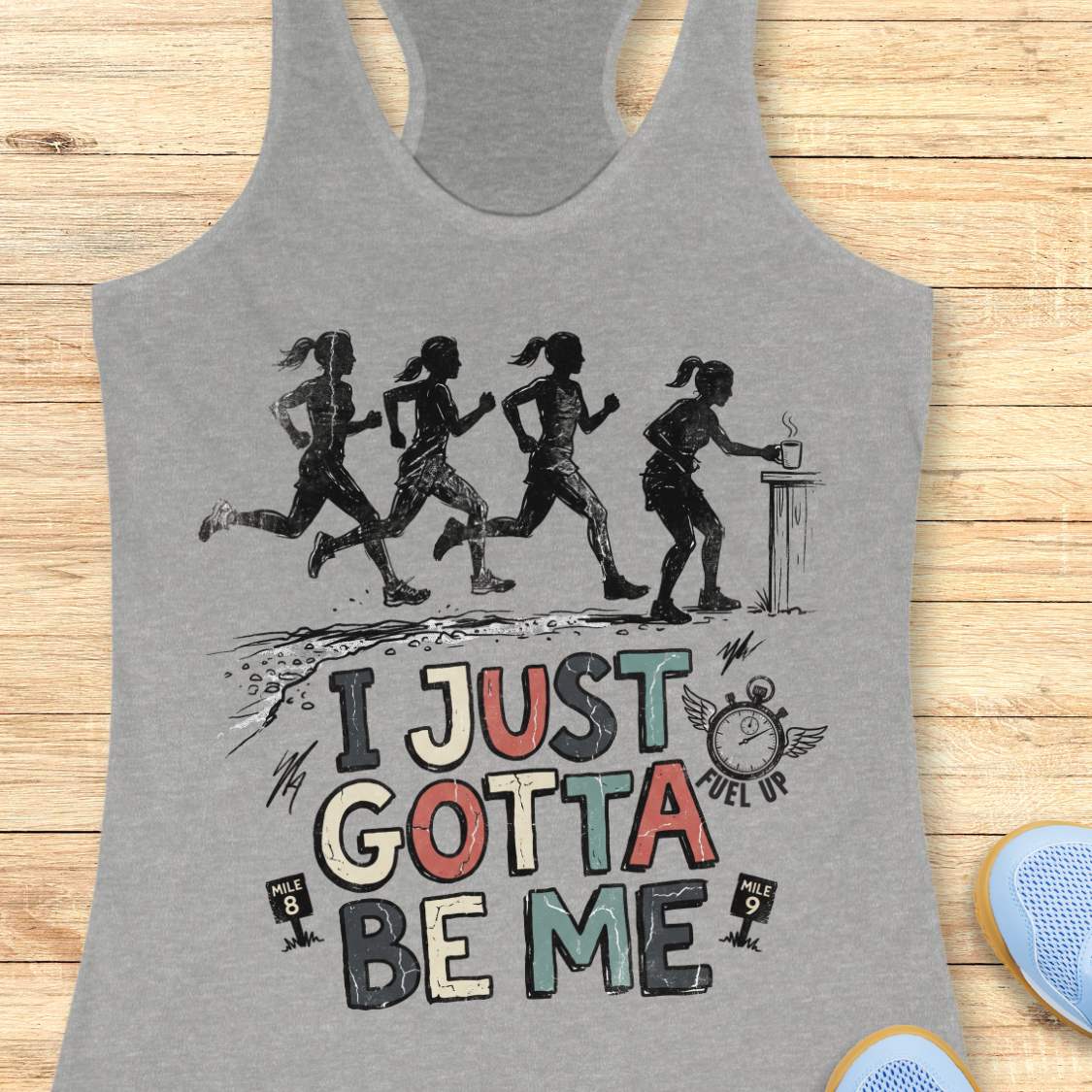 grey racerback tank top featuring a distressed vintage-style running design. Four runners move along a trail, while one runner pauses at a table to grab a steaming cup, suggesting a mid-run coffee or fuel stop. Below the illustration is the phrase “I JUST GOTTA BE ME” in large multicolored lettering, accompanied by a winged stopwatch labeled “Fuel Up” and mile markers reading “Mile 8” and “Mile 9.” The design celebrates runners who embrace their own pace and priorities, including stopping to refuel dur