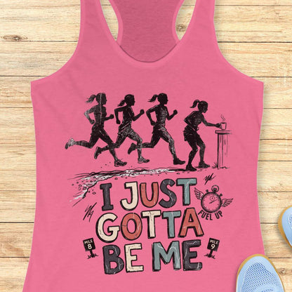 pink racerback tank top featuring a distressed vintage-style running design. Four runners move along a trail, while one runner pauses at a table to grab a steaming cup, suggesting a mid-run coffee or fuel stop. Below the illustration is the phrase “I JUST GOTTA BE ME” in large multicolored lettering, accompanied by a winged stopwatch labeled “Fuel Up” and mile markers reading “Mile 8” and “Mile 9.” The design celebrates runners who embrace their own pace and priorities, including stopping to refuel dur