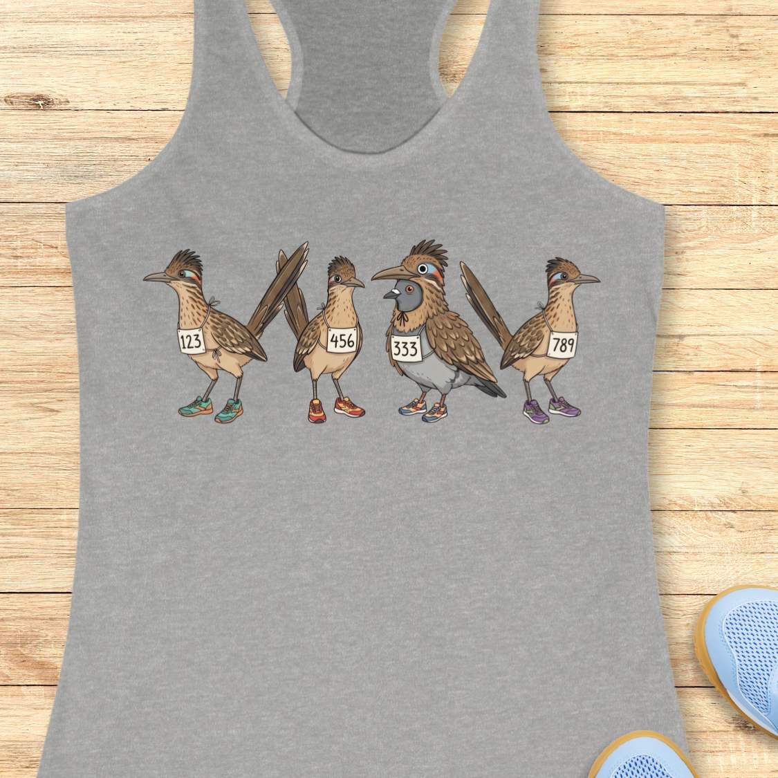 grey racerback tank top featuring a whimsical illustration of four road runner birds wearing race bibs and colorful running shoes. Three road runners stand together while a different bird, a pigeon wearing a road runner costume and race bib, stands among them as part of the group. The playful design symbolizes belonging, inclusion, and the idea that every runner has a place in the running community, regardless of pace, experience, or appearance. The artwork uses soft earth tones with colorful shoe accents 