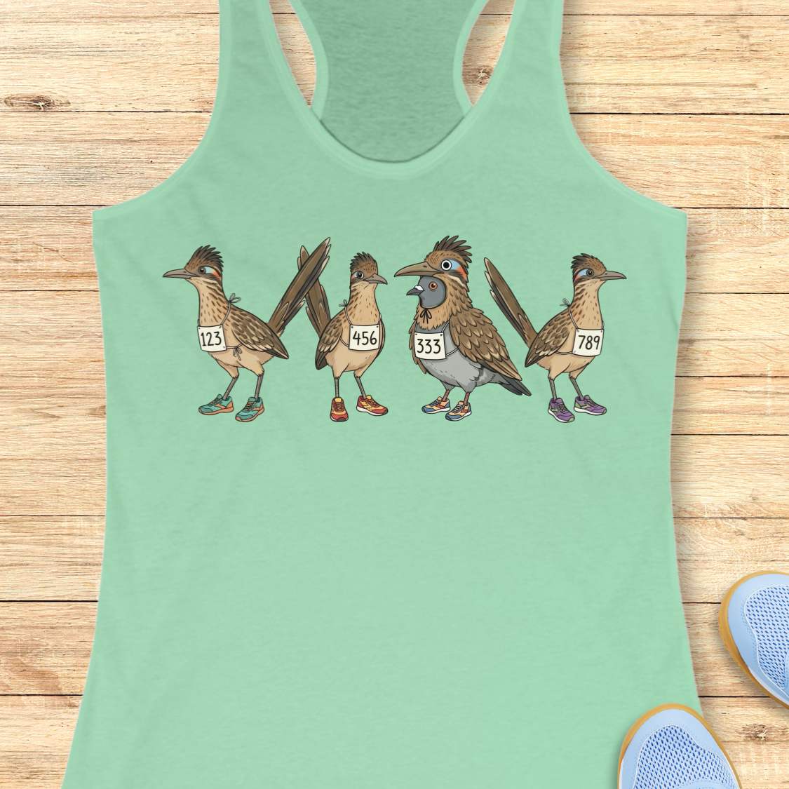 mint racerback tank top featuring a whimsical illustration of four road runner birds wearing race bibs and colorful running shoes. Three road runners stand together while a different bird, a pigeon wearing a road runner costume and race bib, stands among them as part of the group. The playful design symbolizes belonging, inclusion, and the idea that every runner has a place in the running community, regardless of pace, experience, or appearance. The artwork uses soft earth tones with colorful shoe accents 