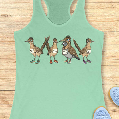 mint racerback tank top featuring a whimsical illustration of four road runner birds wearing race bibs and colorful running shoes. Three road runners stand together while a different bird, a pigeon wearing a road runner costume and race bib, stands among them as part of the group. The playful design symbolizes belonging, inclusion, and the idea that every runner has a place in the running community, regardless of pace, experience, or appearance. The artwork uses soft earth tones with colorful shoe accents 
