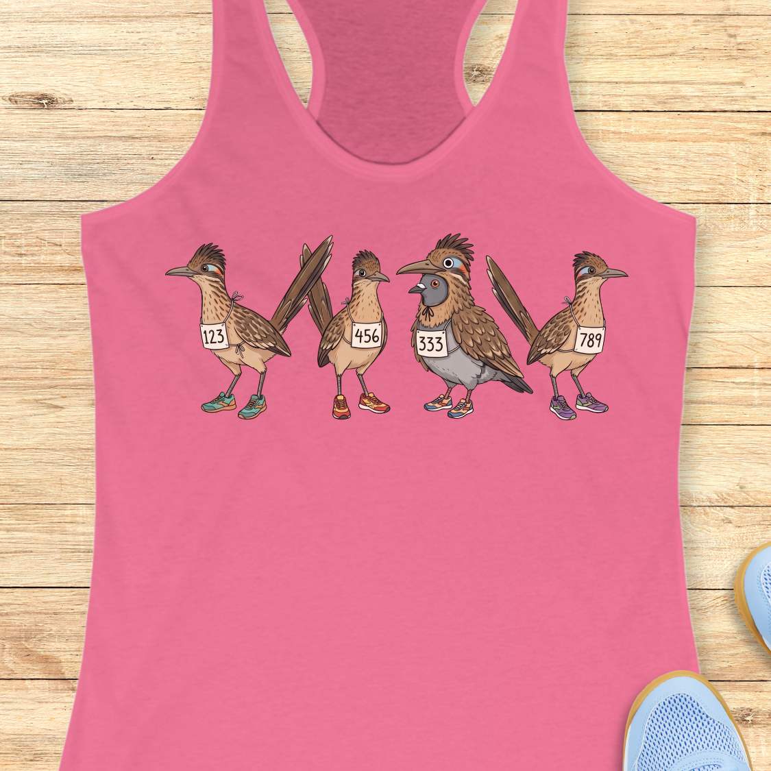 pink racerback tank top featuring a whimsical illustration of four road runner birds wearing race bibs and colorful running shoes. Three road runners stand together while a different bird, a pigeon wearing a road runner costume and race bib, stands among them as part of the group. The playful design symbolizes belonging, inclusion, and the idea that every runner has a place in the running community, regardless of pace, experience, or appearance. The artwork uses soft earth tones with colorful shoe accents 