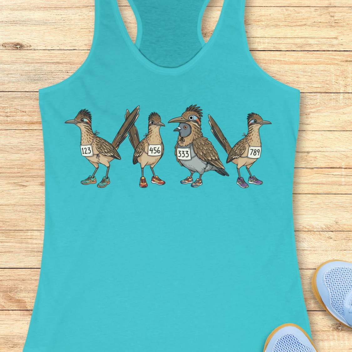 blue racerback tank top featuring a whimsical illustration of four road runner birds wearing race bibs and colorful running shoes. Three road runners stand together while a different bird, a pigeon wearing a road runner costume and race bib, stands among them as part of the group. The playful design symbolizes belonging, inclusion, and the idea that every runner has a place in the running community, regardless of pace, experience, or appearance. The artwork uses soft earth tones with colorful shoe accents 