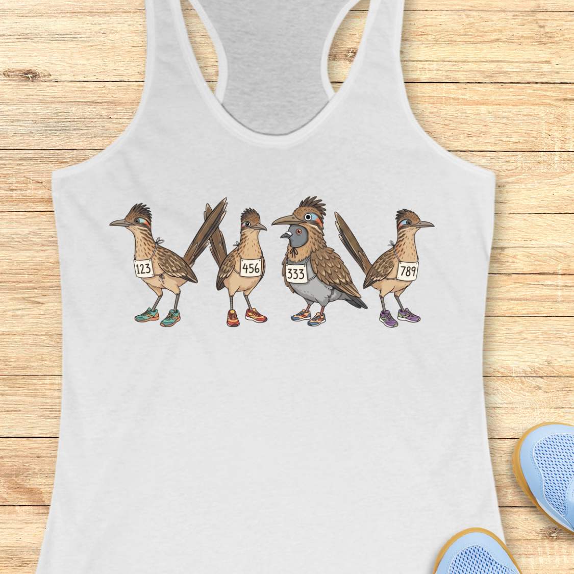White racerback tank top featuring a whimsical illustration of four road runner birds wearing race bibs and colorful running shoes. Three road runners stand together while a different bird, a pigeon wearing a road runner costume and race bib, stands among them as part of the group. The playful design symbolizes belonging, inclusion, and the idea that every runner has a place in the running community, regardless of pace, experience, or appearance. The artwork uses soft earth tones with colorful shoe accents 
