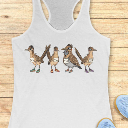 White racerback tank top featuring a whimsical illustration of four road runner birds wearing race bibs and colorful running shoes. Three road runners stand together while a different bird, a pigeon wearing a road runner costume and race bib, stands among them as part of the group. The playful design symbolizes belonging, inclusion, and the idea that every runner has a place in the running community, regardless of pace, experience, or appearance. The artwork uses soft earth tones with colorful shoe accents 