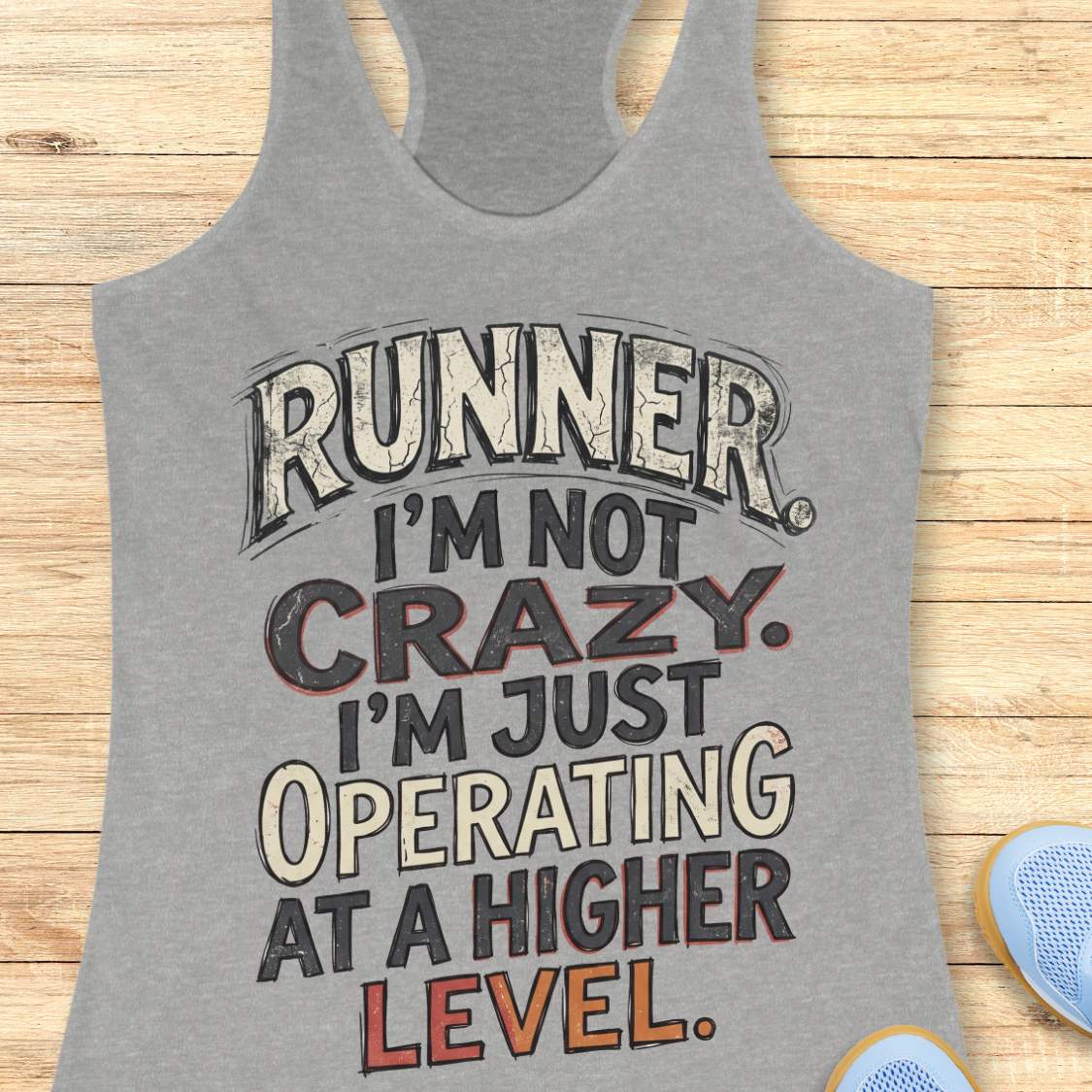 grey racerback tank top featuring bold distressed typography in white, navy blue, and teal. The design displays the humorous runner slogan: “RUNNER. I’M NOT CRAZY. I’M JUST OPERATING AT A HIGHER LEVEL.” The statement is arranged in large stacked lettering with a vintage athletic style, combining confidence, humor, and runner pride. The design celebrates the unique mindset, dedication, and passion of the running community