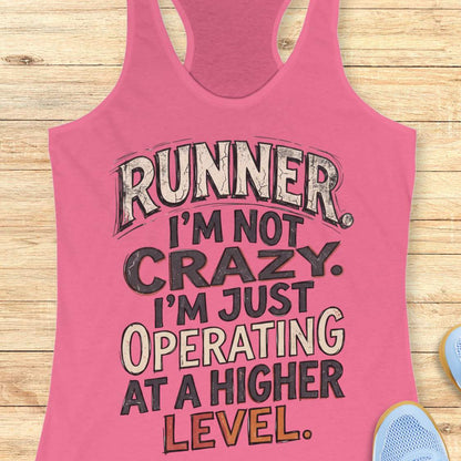 pink racerback tank top featuring bold distressed typography in white, navy blue, and teal. The design displays the humorous runner slogan: “RUNNER. I’M NOT CRAZY. I’M JUST OPERATING AT A HIGHER LEVEL.” The statement is arranged in large stacked lettering with a vintage athletic style, combining confidence, humor, and runner pride. The design celebrates the unique mindset, dedication, and passion of the running community