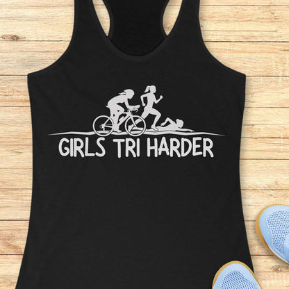Black racerback tank top featuring a clean white graphic celebrating triathlon athletes. The design shows three silhouettes representing the sports of a triathlon: a cyclist riding a bicycle, a runner in motion, and a swimmer moving through the water. Beneath the illustration, bold lettering reads “GIRLS TRI HARDER.” The minimalist design combines athletic pride, determination, and humor, making it a fun statement piece for women who participate in triathlons and endurance sports.