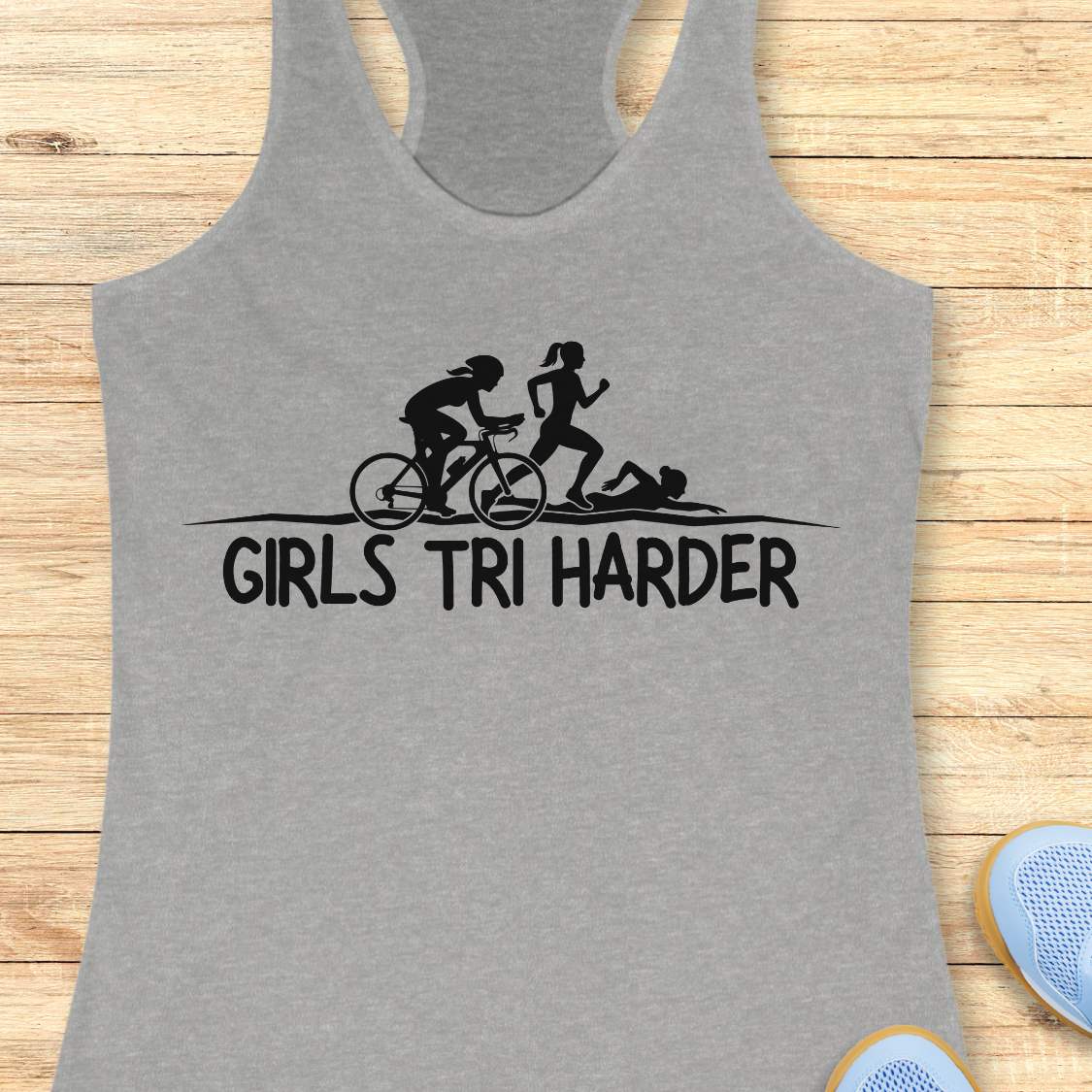 grey racerback tank top featuring a clean white graphic celebrating triathlon athletes. The design shows three silhouettes representing the sports of a triathlon: a cyclist riding a bicycle, a runner in motion, and a swimmer moving through the water. Beneath the illustration, bold lettering reads “GIRLS TRI HARDER.” The minimalist design combines athletic pride, determination, and humor, making it a fun statement piece for women who participate in triathlons and endurance sports.