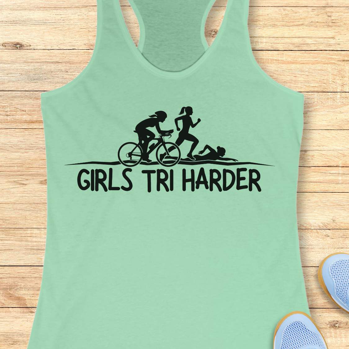 mint racerback tank top featuring a clean white graphic celebrating triathlon athletes. The design shows three silhouettes representing the sports of a triathlon: a cyclist riding a bicycle, a runner in motion, and a swimmer moving through the water. Beneath the illustration, bold lettering reads “GIRLS TRI HARDER.” The minimalist design combines athletic pride, determination, and humor, making it a fun statement piece for women who participate in triathlons and endurance sports.