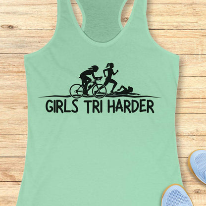 mint racerback tank top featuring a clean white graphic celebrating triathlon athletes. The design shows three silhouettes representing the sports of a triathlon: a cyclist riding a bicycle, a runner in motion, and a swimmer moving through the water. Beneath the illustration, bold lettering reads “GIRLS TRI HARDER.” The minimalist design combines athletic pride, determination, and humor, making it a fun statement piece for women who participate in triathlons and endurance sports.