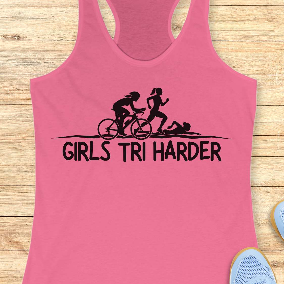pink racerback tank top featuring a clean white graphic celebrating triathlon athletes. The design shows three silhouettes representing the sports of a triathlon: a cyclist riding a bicycle, a runner in motion, and a swimmer moving through the water. Beneath the illustration, bold lettering reads “GIRLS TRI HARDER.” The minimalist design combines athletic pride, determination, and humor, making it a fun statement piece for women who participate in triathlons and endurance sports.