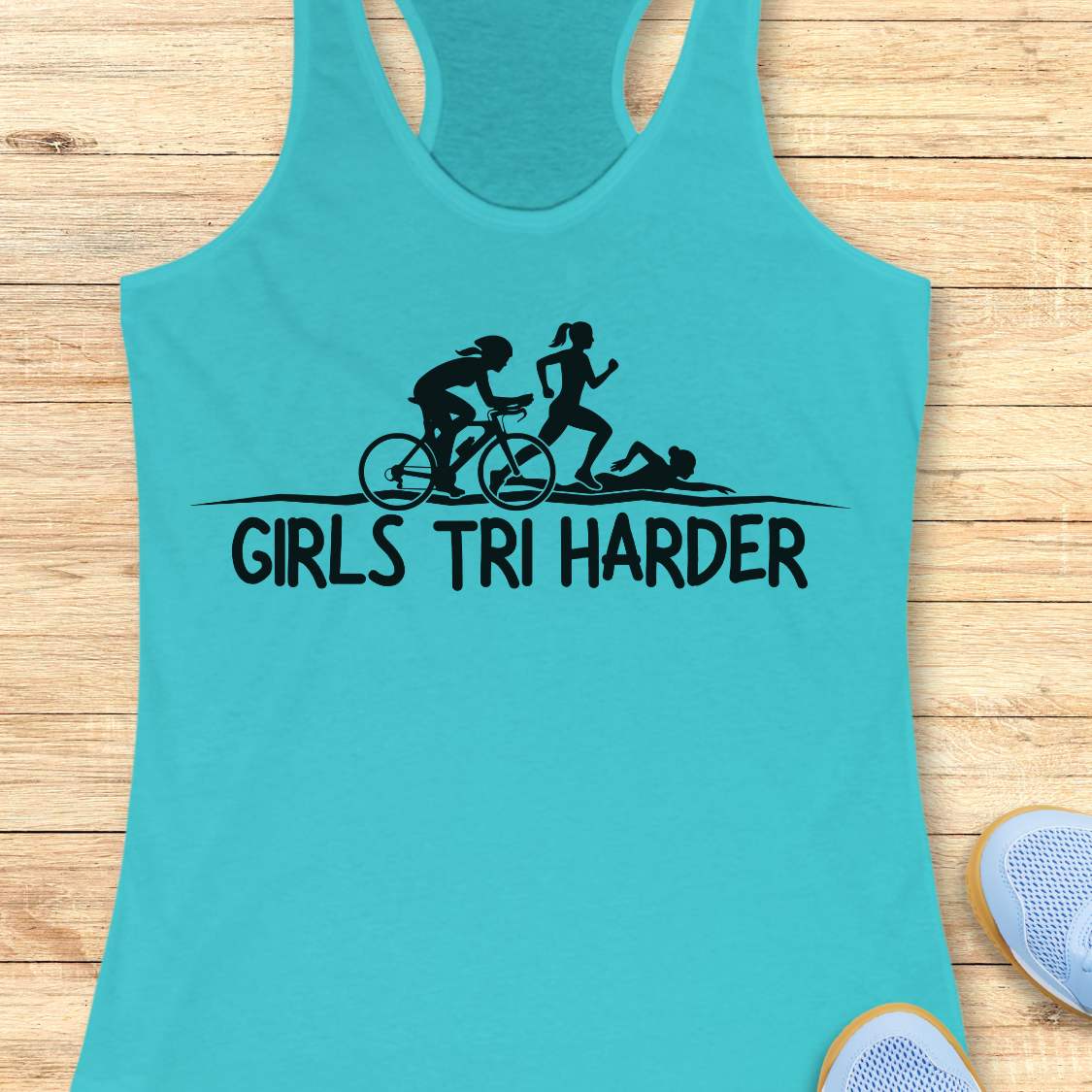 blue racerback tank top featuring a clean white graphic celebrating triathlon athletes. The design shows three silhouettes representing the sports of a triathlon: a cyclist riding a bicycle, a runner in motion, and a swimmer moving through the water. Beneath the illustration, bold lettering reads “GIRLS TRI HARDER.” The minimalist design combines athletic pride, determination, and humor, making it a fun statement piece for women who participate in triathlons and endurance sports.
