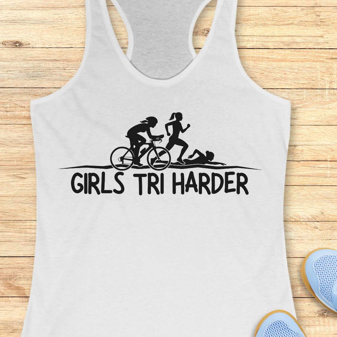 white racerback tank top featuring a clean white graphic celebrating triathlon athletes. The design shows three silhouettes representing the sports of a triathlon: a cyclist riding a bicycle, a runner in motion, and a swimmer moving through the water. Beneath the illustration, bold lettering reads “GIRLS TRI HARDER.” The minimalist design combines athletic pride, determination, and humor, making it a fun statement piece for women who participate in triathlons and endurance sports.