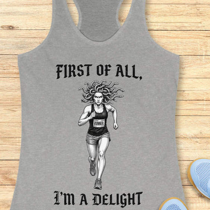 grey women's racerback running tank top displayed on a light wood surface. The design features a black-and-white illustration of a runner with Medusa-style snake hair wearing a race bib. Above the runner, gothic-style text reads “FIRST OF ALL,” and below the runner, matching text reads “I’M A DELIGHT.” Running shoes are partially visible in the corners of the image.