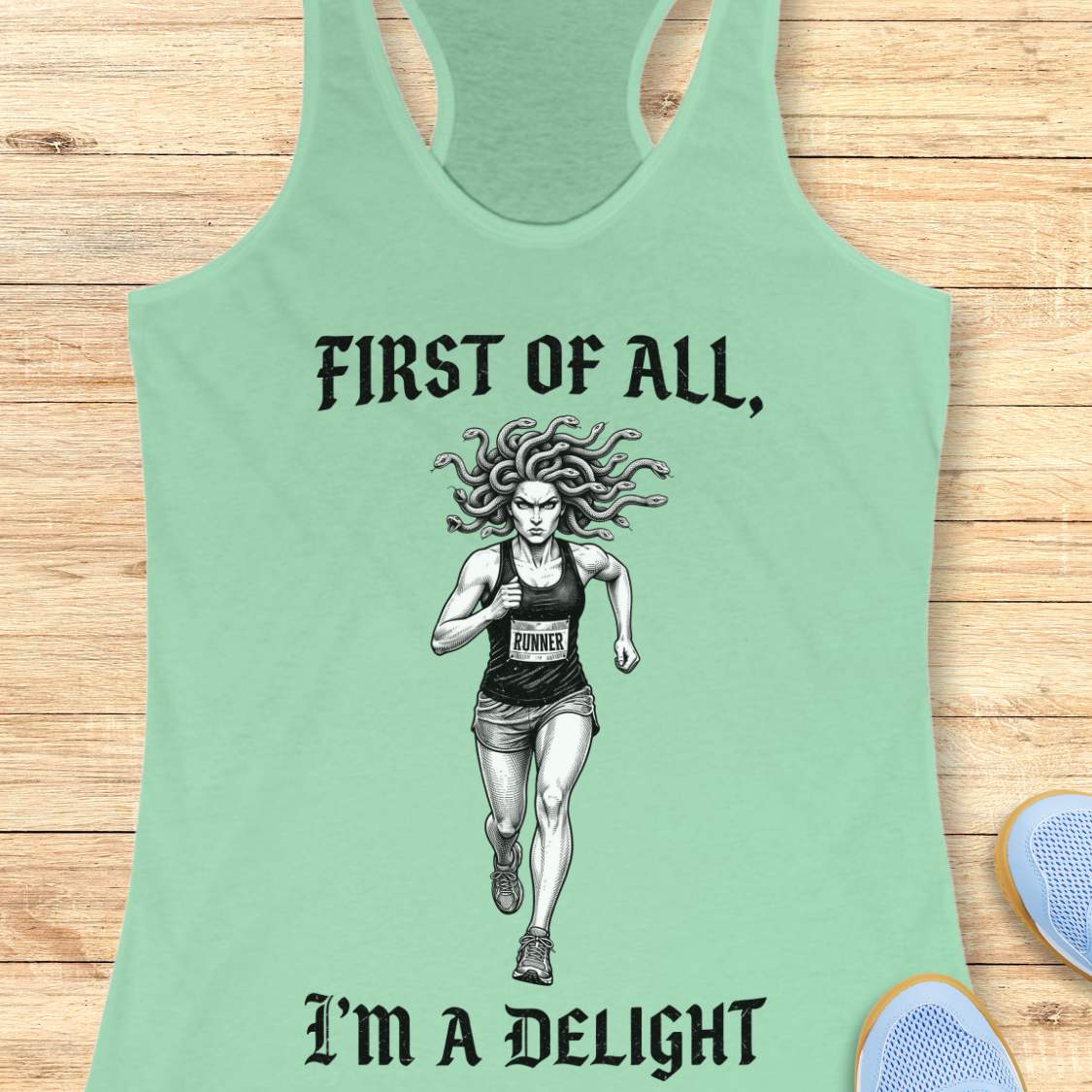 A mint green racerback tank top laid flat on a light wood background, featuring a black graphic of a female runner with snakes in her hair and the text 'FIRST OF ALL, I'M A DELIGHT'.