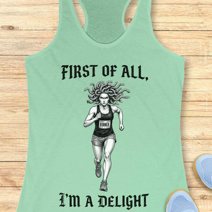 A mint green racerback tank top laid flat on a light wood background, featuring a black graphic of a female runner with snakes in her hair and the text 'FIRST OF ALL, I'M A DELIGHT'.