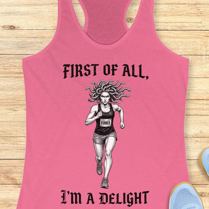 pink women's racerback running tank top displayed on a light wood surface. The design features a black-and-white illustration of a runner with Medusa-style snake hair wearing a race bib. Above the runner, gothic-style text reads “FIRST OF ALL,” and below the runner, matching text reads “I’M A DELIGHT.” Running shoes are partially visible in the corners of the image.