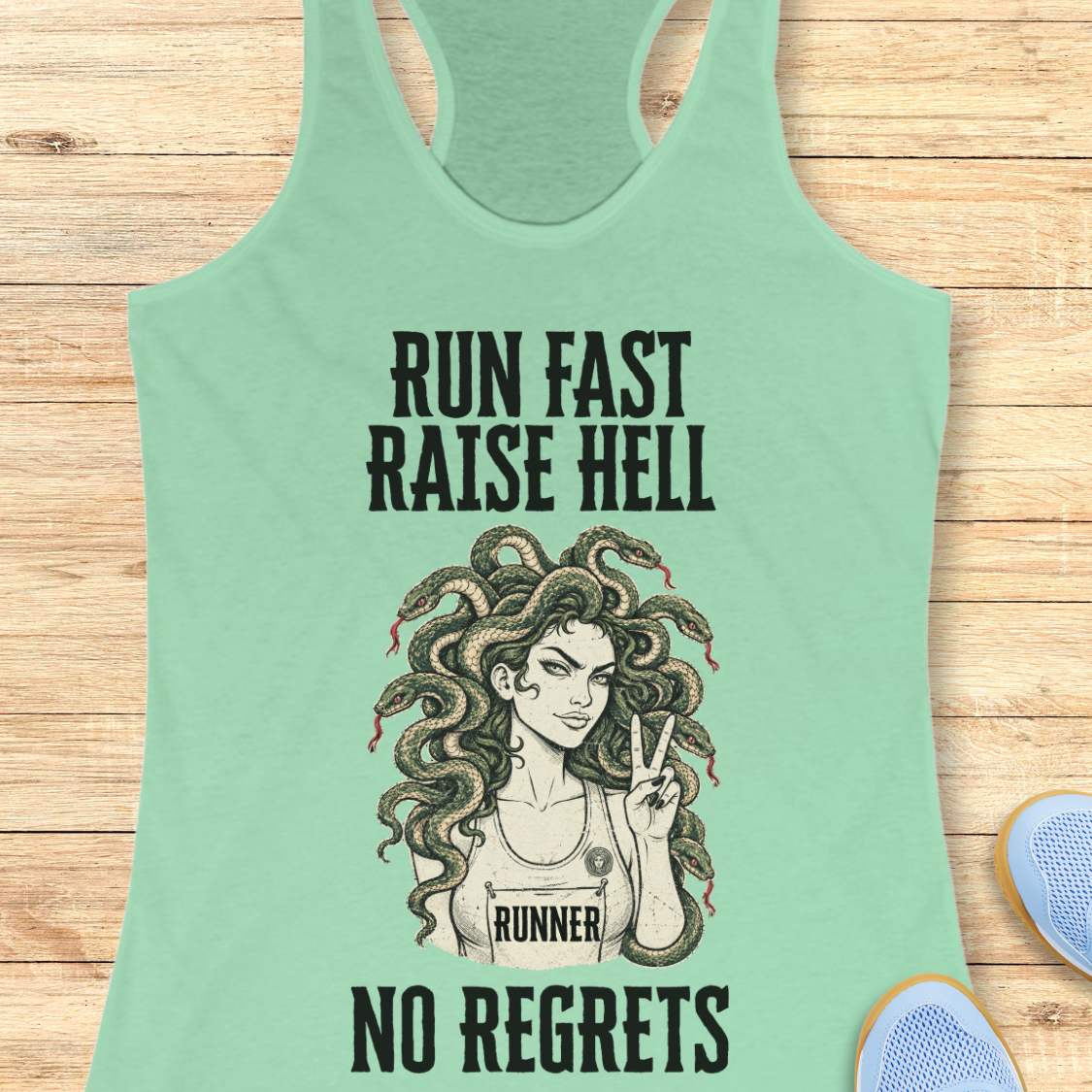 A mint green racerback tank top with a graphic of a Medusa-style runner and the text 'RUN FAST RAISE HELL NO REGRETS' in black, laid flat on a light wood background with partial blue shoes visible at the edges.