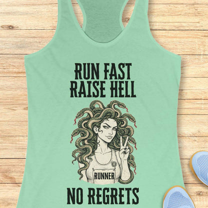 A mint green racerback tank top with a graphic of a Medusa-style runner and the text 'RUN FAST RAISE HELL NO REGRETS' in black, laid flat on a light wood background with partial blue shoes visible at the edges.