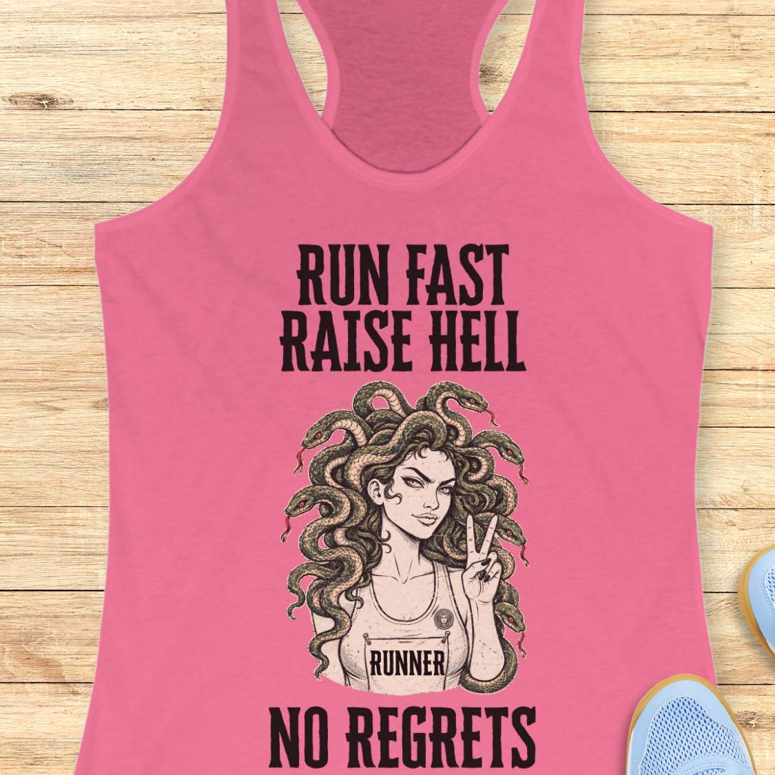 A pink racerback tank top laid flat on a light wood background, featuring a black graphic of a woman with Medusa-style hair and the text 'RUN FAST RAISE HELL NO REGRETS' with 'RUNNER' on the chest. Partial blue shoes are visible at the edges.