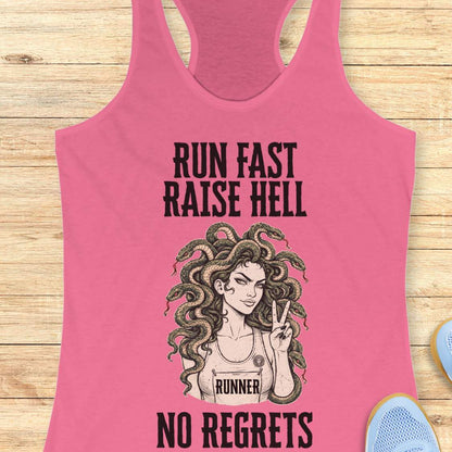 A pink racerback tank top laid flat on a light wood background, featuring a black graphic of a woman with Medusa-style hair and the text 'RUN FAST RAISE HELL NO REGRETS' with 'RUNNER' on the chest. Partial blue shoes are visible at the edges.