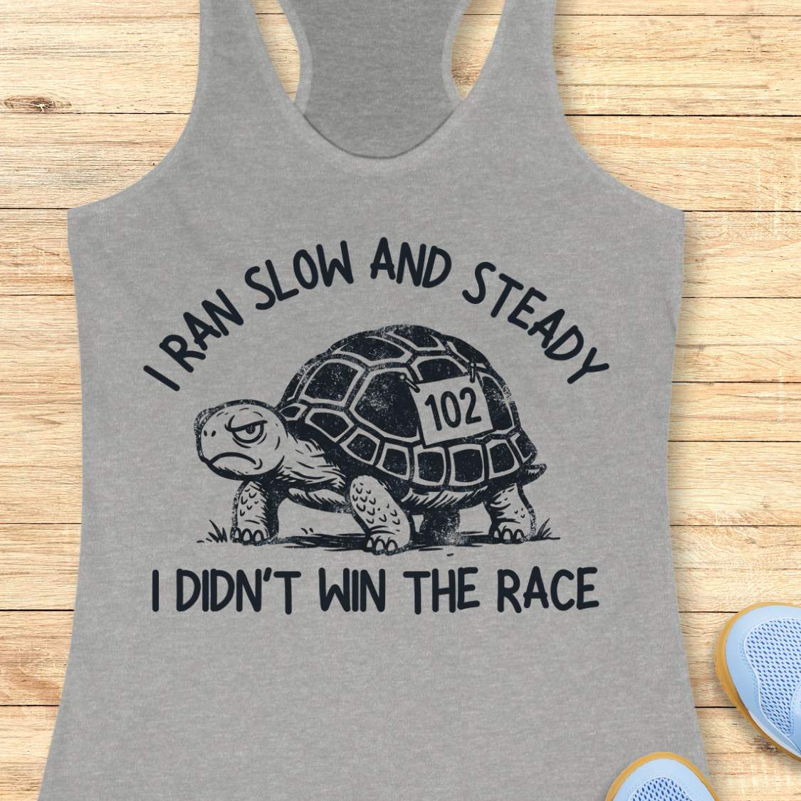 A heather gray racerback tank top laid flat on a light wood surface, featuring a black graphic of a turtle with the number 102 on its shell and the text 'I RAN SLOW AND STEADY I DIDN'T WIN THE RACE'.