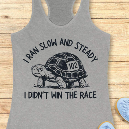 A heather gray racerback tank top laid flat on a light wood surface, featuring a black graphic of a turtle with the number 102 on its shell and the text 'I RAN SLOW AND STEADY I DIDN'T WIN THE RACE'.