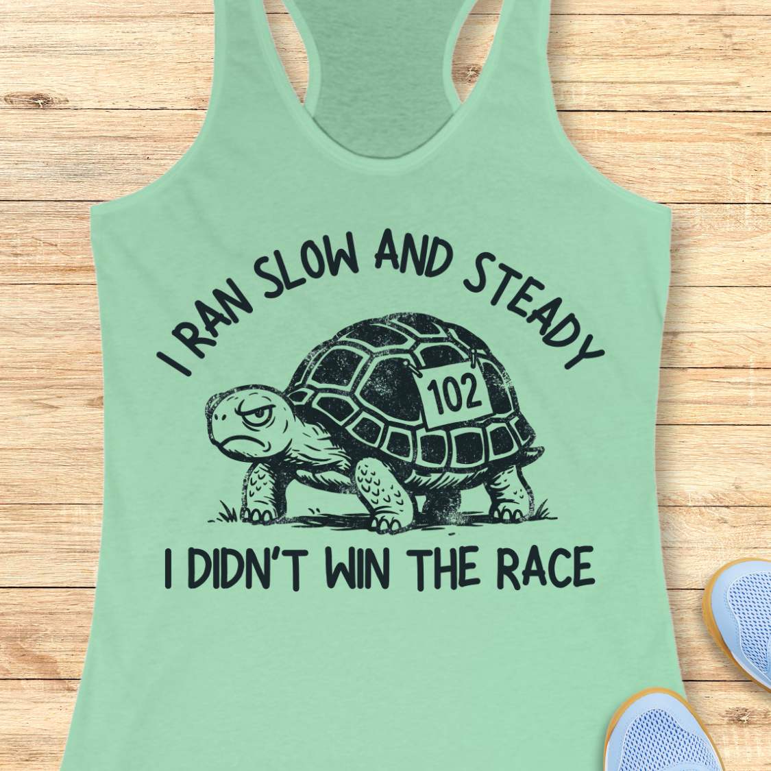 A mint green racerback tank top laid flat on a light wood background, featuring a black graphic of a tortoise with the number 102 and the text 'I RAN SLOW AND STEADY I DIDN'T WIN THE RACE'.