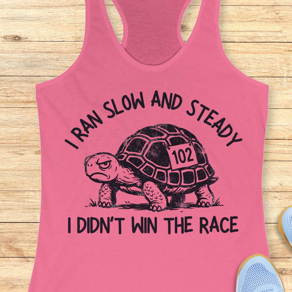 A pink racerback tank top laid flat on a light wood background, featuring a black graphic of a turtle with the number 102 and the text 'I RAN SLOW AND STEADY I DIDN'T WIN THE RACE'.