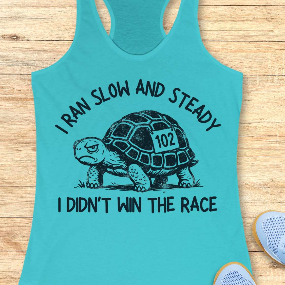 Turquoise women's racerback running tank top displayed on a light wood background. The design features a black illustrated tortoise wearing a race bib numbered “102.” Curved text above the tortoise reads “I RAN SLOW AND STEADY,” and text below reads “I DIDN’T WIN THE RACE.” Partial running shoes are visible in the lower corners of the image.