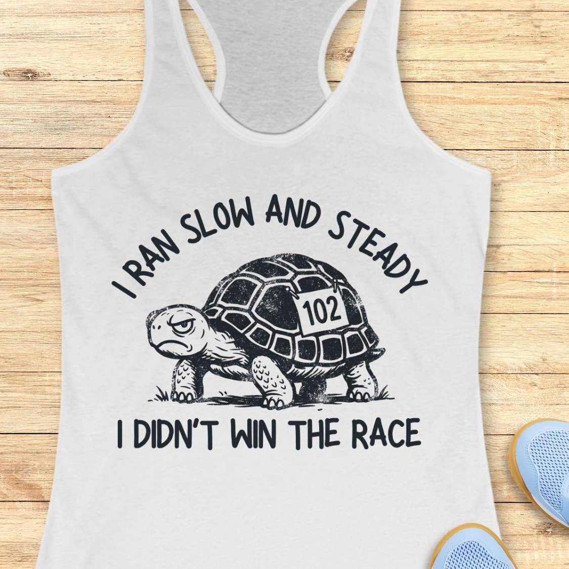 A white racerback tank top laid flat on a light wood background, featuring a black graphic of a tortoise with the number 102 on its shell and the text 'I RAN SLOW AND STEADY I DIDN'T WIN THE RACE'. Partial blue shoes are visible at the bottom right edge.