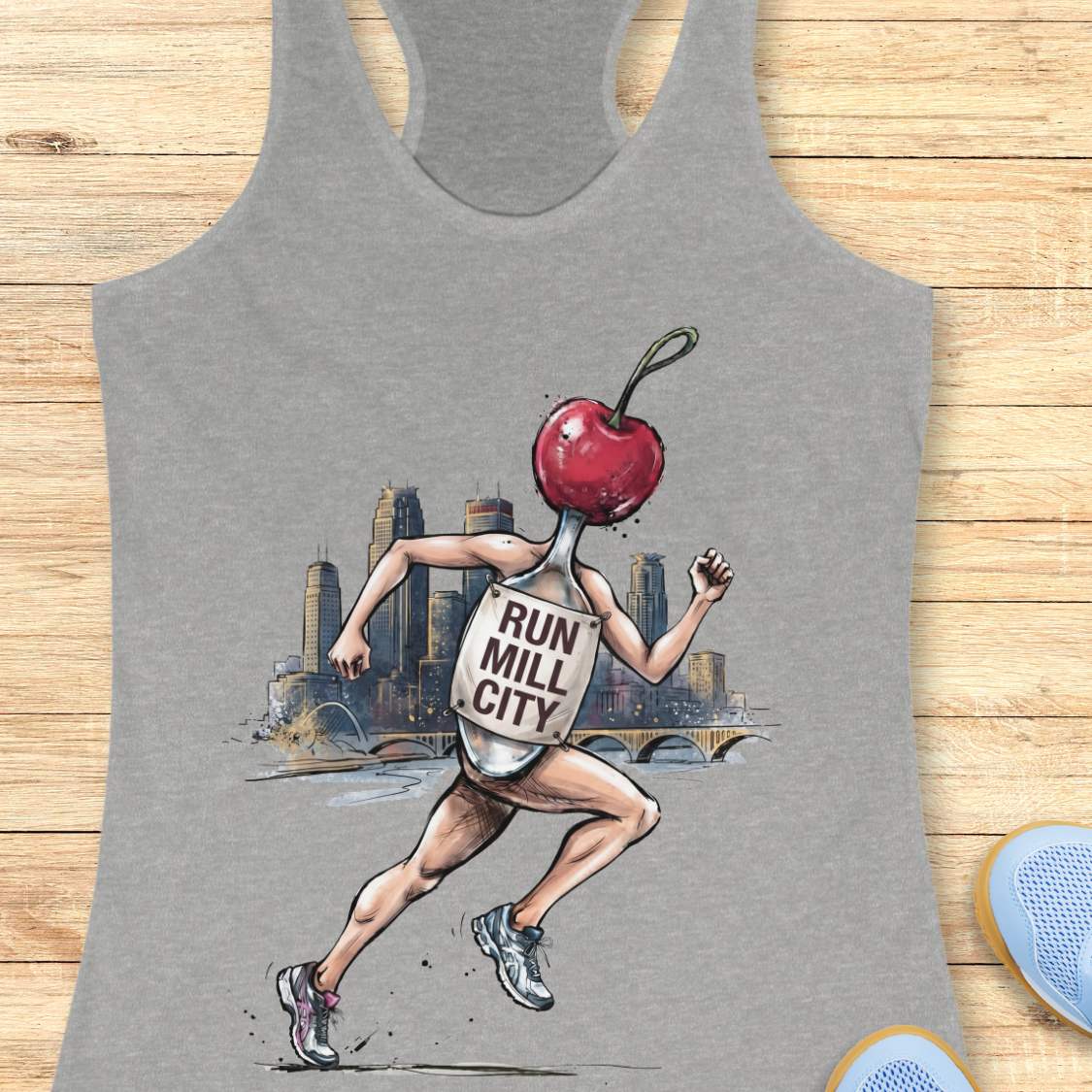 A heather gray racerback tank top laid flat on a light wood surface, featuring a large graphic print of a runner with a red cherry for a head and a milk bottle labeled 'RUN MILL CITY' on their back. Partial blue shoes are visible at the bottom right edge.