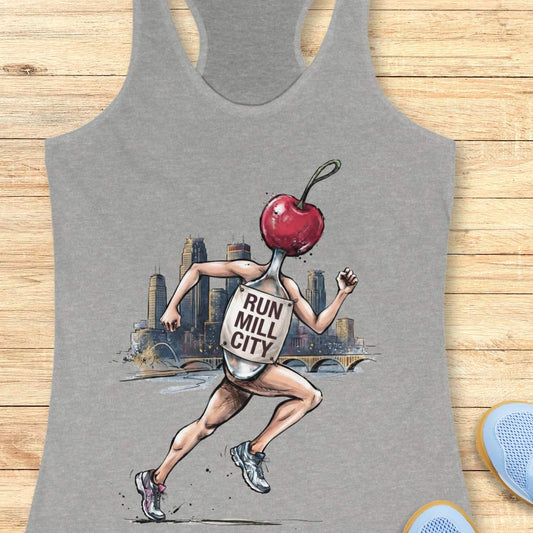 A heather gray racerback tank top laid flat on a light wood surface, featuring a large graphic print of a runner with a red cherry for a head and a milk bottle labeled 'RUN MILL CITY' on their back. Partial blue shoes are visible at the bottom right edge.