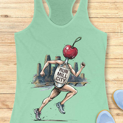 A mint green racerback tank top laid flat on a light wood plank background. The front of the tank top features a graphic illustration of a running figure with a red cherry for a head, wearing a tank top that reads 'RUN MILL CITY.